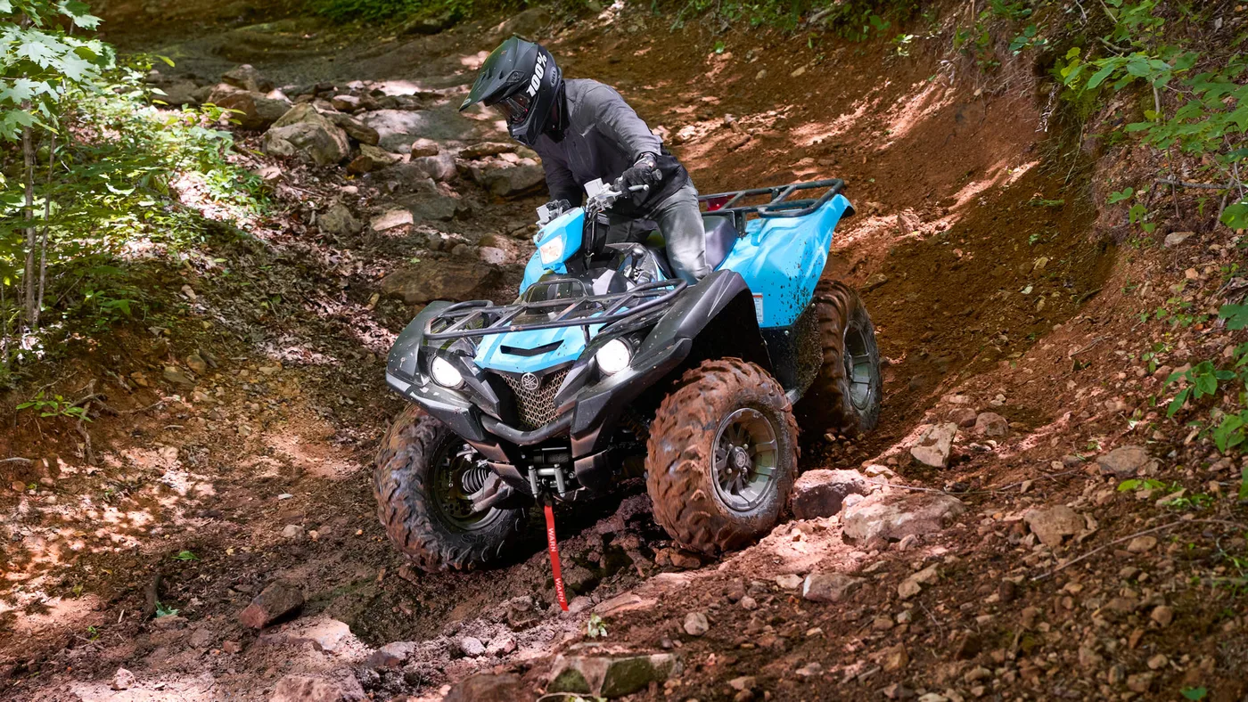 Rider on a blue quad crossing a rocky woodland trail, as featured on QuadParts